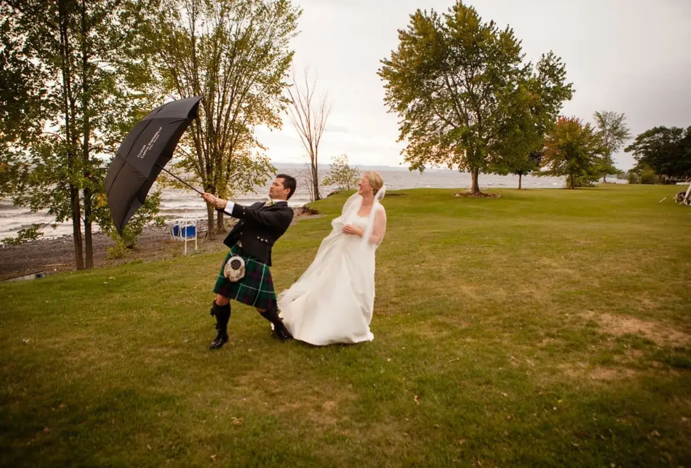 A bride and groom holding an umbrella in the grass during their offsite wedding ceremony.