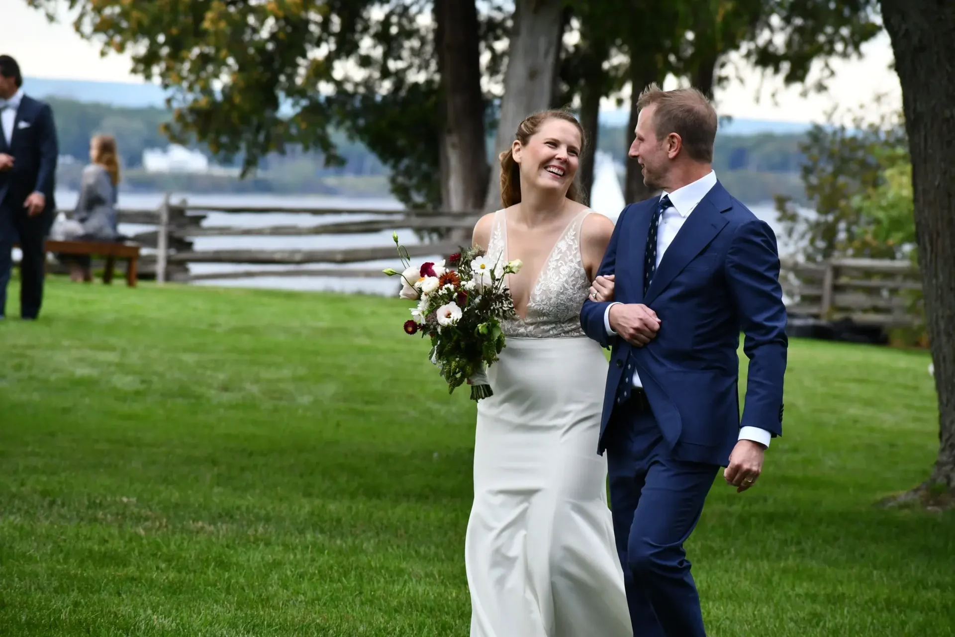 A bride and groom enjoy their offsite wedding ceremony, walking hand in hand through a picturesque grassy area near a serene lake.