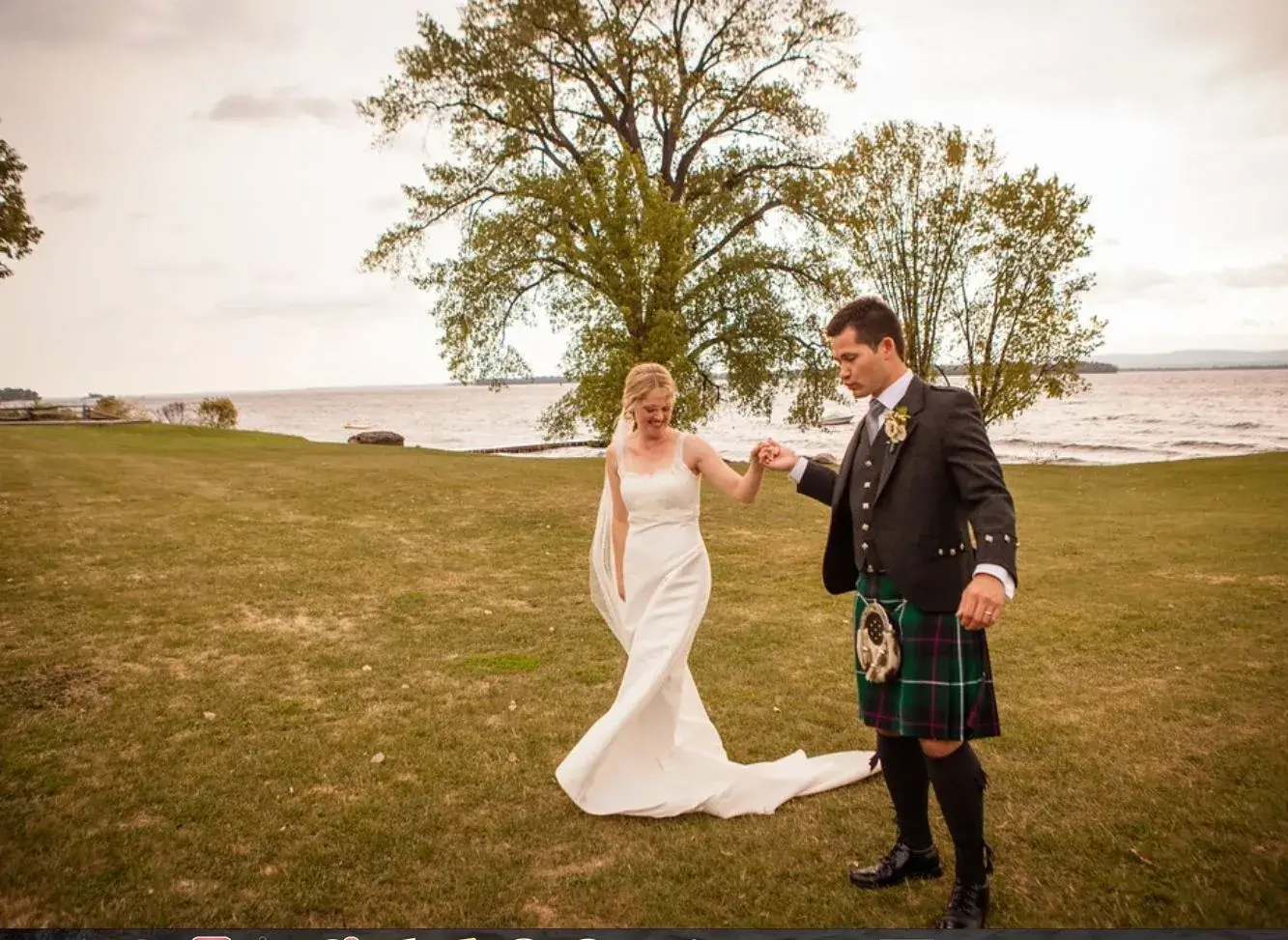 A bride and groom in kilts walking in front of a lake during their offsite wedding.