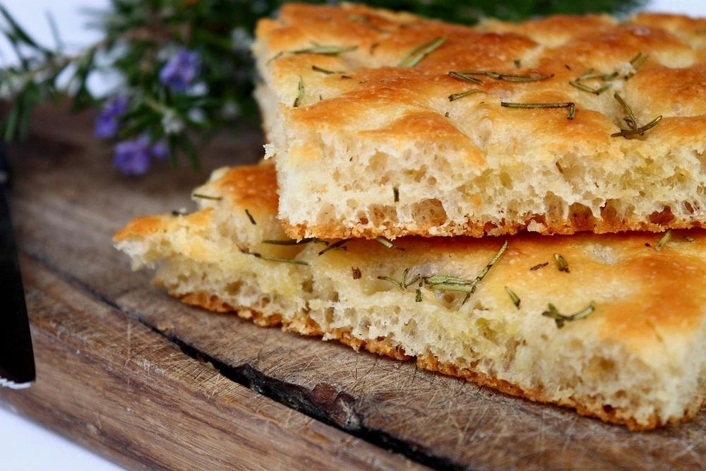 A slice of House Made Focaccia (daily) with rosemary and thyme on a cutting board.