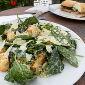 A plate of Shaved Kale & Brussels Sprout Caesar and croutons on a table next to a sandwich.