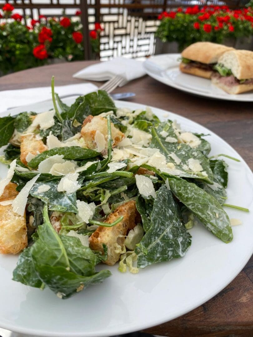 A plate of Shaved Kale & Brussels Sprout Caesar and croutons on a table next to a sandwich.