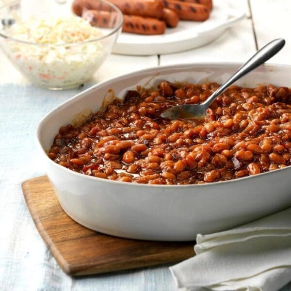 A white dish with New England Baked Beans and a spoon on a wooden cutting board.